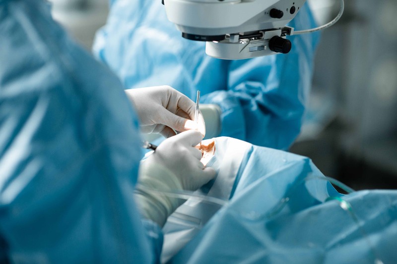 Schräger Blick auf die Hände des Chirurgen während der grauen Star Operation. Das Gesicht des Patientin ist mit einem blauen Tuch abgedeckt, der Chirurg trägt weiße Handschuhe und hält ein silberfarbenes Instrument in der Hand / © Till Findl